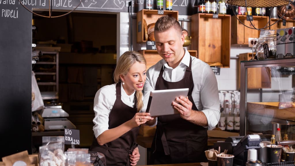 Two café employees in aprons look at a tablet behind the counter, surrounded by shelves and coffee products, as they discuss the California transfer liquor license process to expand their beverage options.